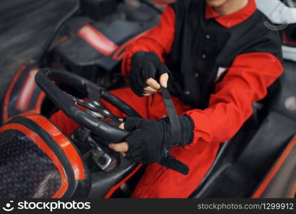 Kart racer in red uniform puts on gloves, karting auto sport indoor. Speed race on close go-kart track with tire barrier. Fast vehicle competition, high adrenaline hobby. Kart racer in red uniform puts on gloves