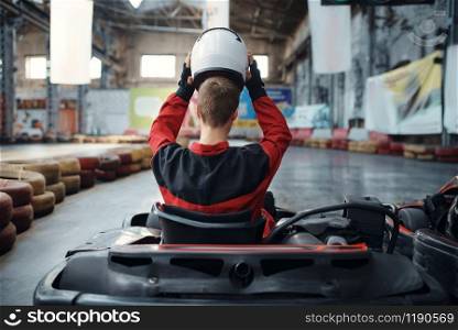 Kart racer puts on helmet, back view, karting indoor. Speed race on close go-cart track with tire barrier. Fast vehicle competition, high adrenaline leisure