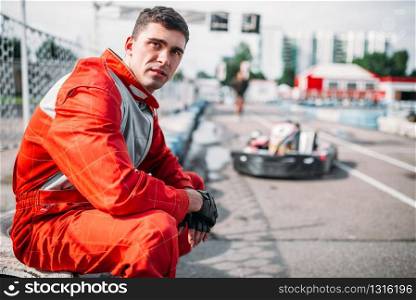Karting racer sits on a tire, kart on background, outdoor track. Motor sport, carting race
