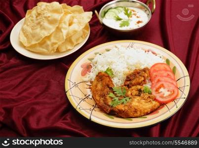 Kashmiri chicken in a serving bowl beside a plate of pappadums and a bowl of raita. The chicken is marinated with tomato and spice paste and the fried with ginger and garlic.