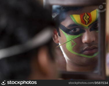 Kathakali dancer looking in the mirror and applying green colour on his face. 