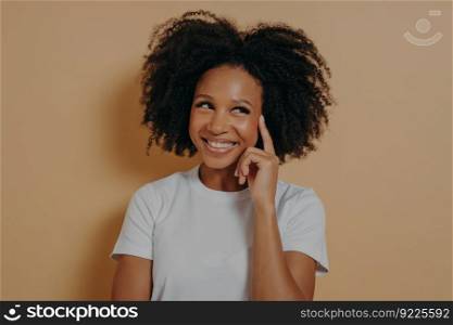 Keep it in mind. Portrait of young positive pensive african woman with curly hair touching temple with forefinger and smiling cheerfully, having good idea, posing isolated over beige background. Pensive african woman with curly hair touching temple with forefinger, isolated on beige background