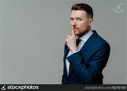Keep it in secret. Young businessman in formal wear suit showing shh sign, keeping finger near mouth while standing against grey studio background, looking at camera with shushing expression. Young businessman in suit asking to keep information in secret