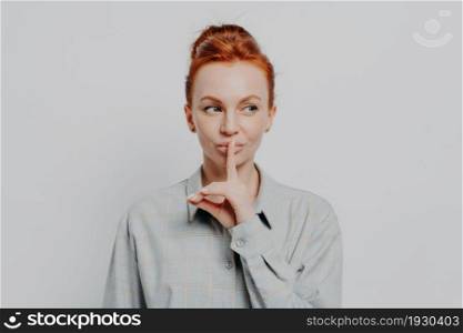 Keep it in secret. Young ginger woman with hair in bun showing silence sign, keeping finger on lips and looking aside at copy space while posing isolated on grey studio background. Privacy concept. Young ginger woman keeping finger on lips and looking aside while standing against grey wall