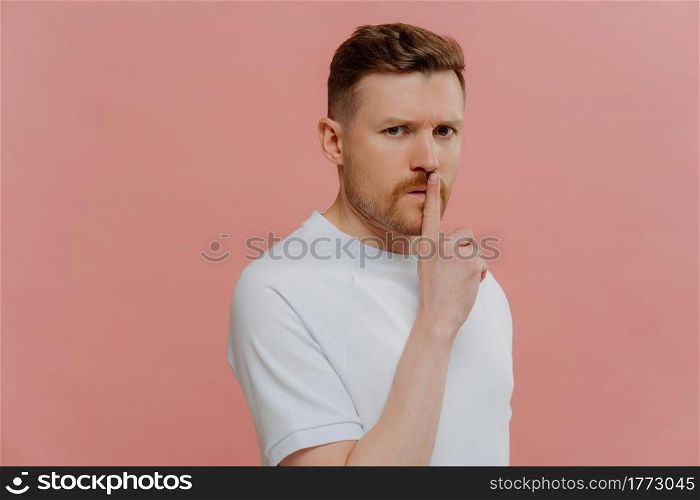 Keep it in secret. Young unshaven man in white tshirt showing silence gesture, making shh and looking at camera with mysterious face expression while standing against pink studio background. Young unshaven mysterious man in white shirt asking to keep information in secret