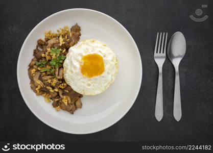 Khao Moo Tod Kratiam Kai Dao, Thai food, streamed rice topped with fried pork with garlic and fried egg in ceramic plate beside fork and spoon on dark tone texture background