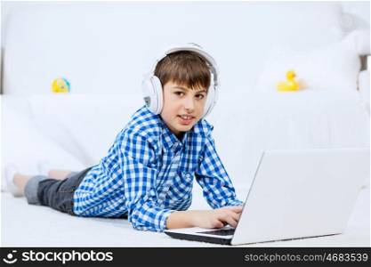Kid enjoying leisure time. Boy of school age listening music from laptop through headphones while lying on floor