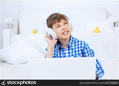 Kid enjoying leisure time. Boy of school age listening music from laptop through headphones while sitting on floor
