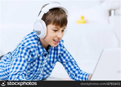 Kid enjoying leisure time. Boy of school age listening music from laptop through headphones while lying on floor