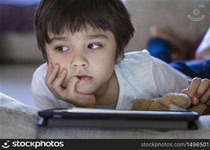 Kid sad face looking deep in thought, Bored Child boy lying on sofa playing game on tablet ,Home schooling,Social Distance,E-learning online education