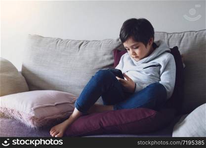 Kid self isolation using cellphone for homework, Sad child sitting on sofa looking down deep in thought,Boy learning on internet from home during school of. Home schooling, learning online education