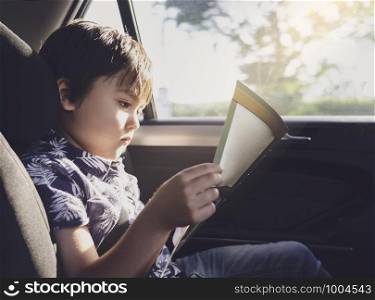 Kid siting on car seat and reading a book, Little boy sitting in the car in child safety seat, Portrait of toddler entertaining himserf on a road trip. Concept of safety taveling by car with children