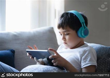 kid sitting on sofa watching cartoon on tablet, Cute boy sitting next to window playing game online with friends,