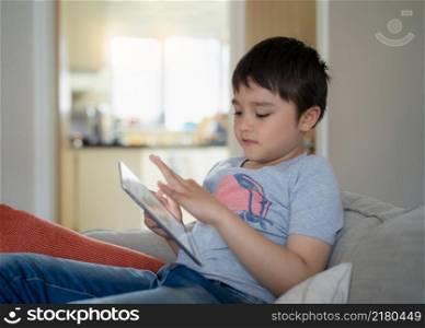 Kid sitting on sofa watching cartoon or playing game on tablet, Child boy using digital pad learning lesson online on internet, Home schooling,Distance learning online education concep