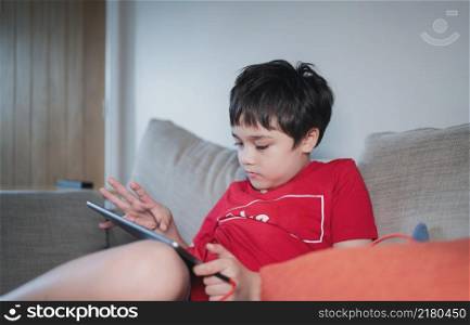 Kid sitting on sofa watching cartoon or playing game on tablet, Child boy using digital pad learning lesson online on internet, Home schooling,Distance learning online education concept