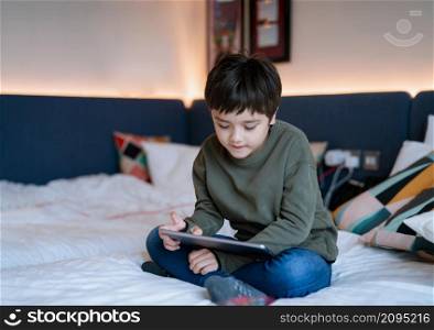 Kid watching cartoon and chatting with friends on tablet, Happy boy sitting in bed playing games online on digital pad, Portrait Child relaxing at home in his bed room on weekend.