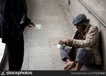 Kind Businessman hold dollar bill money to donate to old beggar or homeless man at city walk in urban town. Poverty and social issue concept. Give and share during covid-19 or coronavirus.