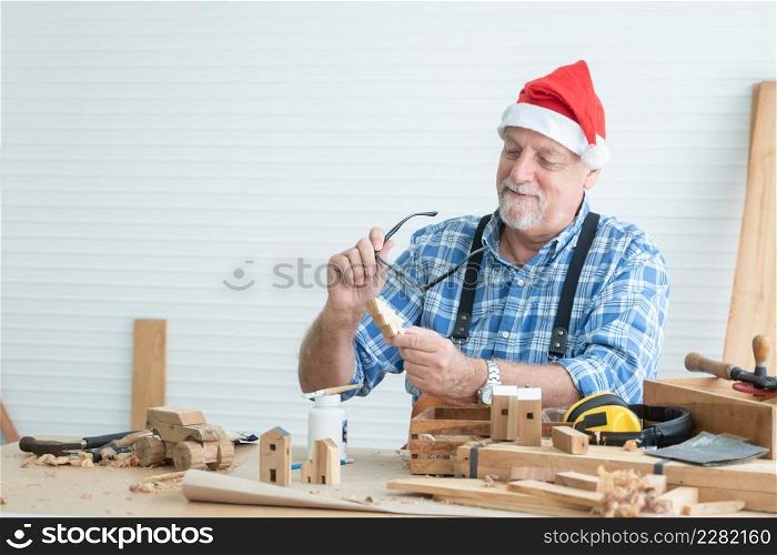 Kind elderly Caucasian carpenter wearing Santa hat working on DIY wood at home. Senior man holding eye glasses and small wooden Christmas tree while producing wooden toys for gifts in holiday season