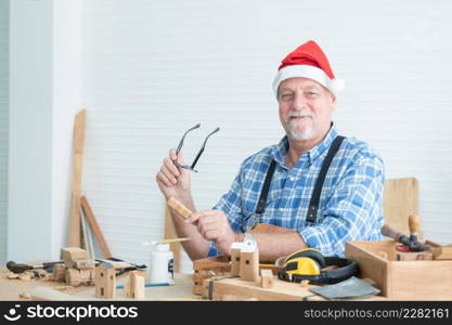 Kind elderly Caucasian carpenter wearing Santa hat working on DIY wood at home. Senior man holding eye glasses and small wooden Christmas tree while producing wooden toys for gifts in holiday season