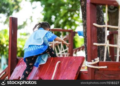 Kindergarten child girl climb up from wooden slope of kid playground with green foliage bokeh background. Outdoors wood equipment for children game.