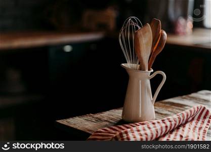 Kitchen accessories on wooden table. Utensils in white ceramic jar against dark background. Rustic style. Dishware for preparing meal. Wooden spoons shpatula and whisk