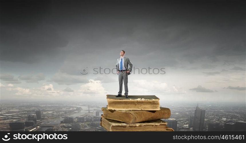 Knowledge and education are his advantage. Young confident businessman with arms on waist standing on pile of old books