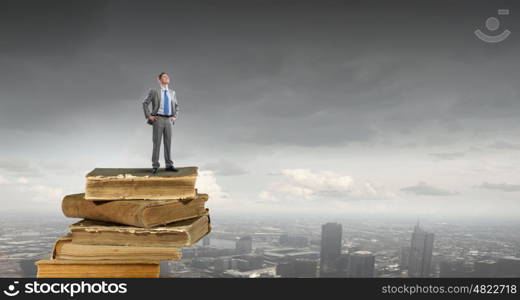 Knowledge and education are his advantage. Young confident businessman with arms on waist standing on pile of old books