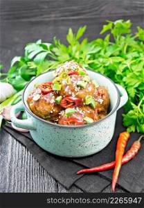 Korean meatballs in sauce of starch, soy sauce, vinegar and apricot jam, sprinkled with green onions, hot peppers and sesame seeds in a saucepan on a napkin against wooden board background