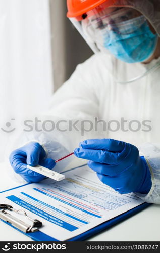 Lab technician medical scientist performing Coronavirus COVID-19 rapid diagnostic testing for antibodies, wearing personal protective equipment PPE, placing blood sample specimen on test using pipette