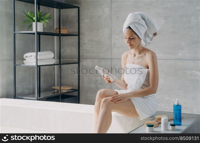 Lady applying body lotion from bottle to her leg after bathing. Attractive european girl wrapped in towel. Young woman takes shower in morning at home. Anti-cellulite massage, hygiene and bodycare.. Young lady applying body lotion from bottle to her leg after bathing. Anti-cellulite massage.