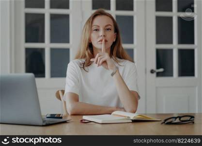 Lady gestures hush. Young businesswoman is working from home. Tender caucasian woman in white t-shirt is freelancer, entrepreneur or accountant having remote work. Concept of secret and privacy.. Lady gestures hush in concept of secret and privacy. Young businesswoman is working from home.
