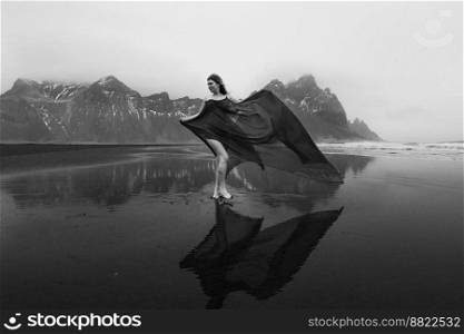 Lady in waving chiffon dress on empty beach monochrome scenic photography. Picture of person with hills on background. High quality wallpaper. Photo concept for ads, travel blog, magazine, article. Lady in waving chiffon dress on empty beach monochrome scenic photography