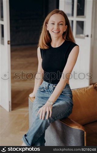 Lady is sitting on an armchair armrest and smiling. Portrait of gorgeous slim blonde young model. Redhead lovely lady in casual outfit. Happy european woman in luxurious apartment.. Lady is sitting on an armchair armrest and smiling. Portrait of gorgeous slim blonde young model.
