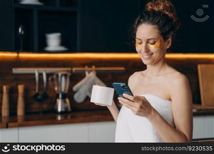 Lady relaxing at home, chatting on phone and having tea. Happy hispanic girl wrapped in towel after spa procedures. Young woman applies anti wrinkle eye patches. Body care, beauty and wellness.. Lady relaxing at home, chatting on phone and having tea. Hispanic girl wrapped in towel after spa.