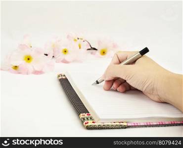 Lady's hand is writing diary with pink artificial flower over white background