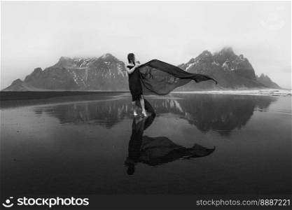 Lady with red waving fabric on northern beach monochrome scenic photography. Picture of person with hills on background. High quality wallpaper. Photo concept for ads, travel blog, magazine, article. Lady with red waving fabric on northern beach monochrome scenic photography