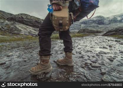 Lady with rucksack walking across shallow creek scenic photography. Picture of person with mountains on background. High quality wallpaper. Photo concept for ads, travel blog, magazine, article. Lady with rucksack walking across shallow creek scenic photography