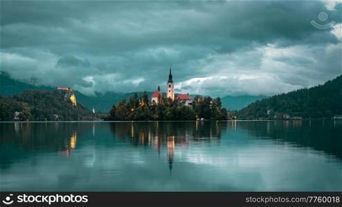 Lake Bled in Slovenia. Sunset view on Assumption of Maria Church against cloudy Julian Alpine mountains. Church reflecting in lake Bled waters.. Lake Bled, view on cloudy church