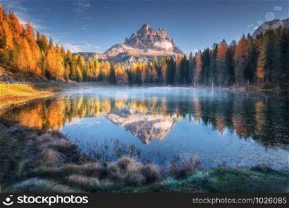 Lake with reflection of mountains at sunrise in autumn in Dolomites, Italy. Landscape with Antorno lake, blue fog over the water, trees with orange leaves and high rocks in fall. Colorful forest. Lake in fog with reflection of mountains at sunrise in autumn