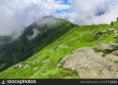 Landscape of a trekker walking at the Chanderkhani pass, India, a very huge green grass covered mountain at an altitude of 12000 ft, where the sky is bluer and clouds are below as well as above.