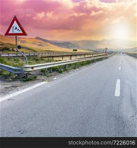 "Landscape of Sicilian hills at sunrise. Asphalt road elevated by columns due of frequent earthquakes of the island of Sicily. Road sign: "Side wind""
