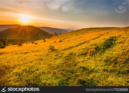 Landscape sunset in mountains with forest, green grass and big shining sun on dramatic sky