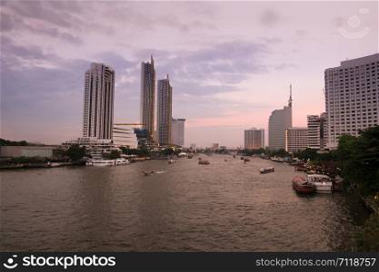 Landscape view of sunset at Chao Phraya river with a view of boats and modern buildings along the riverside. Bangkok, Thailand.