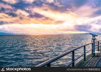 Landscape with a colorful sunset over the Bodensee lake and its shore, equipped with tourists binoculars and metal railing for safety.