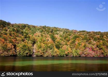Landscape with early autumn forest on a hill, lake and blue sky