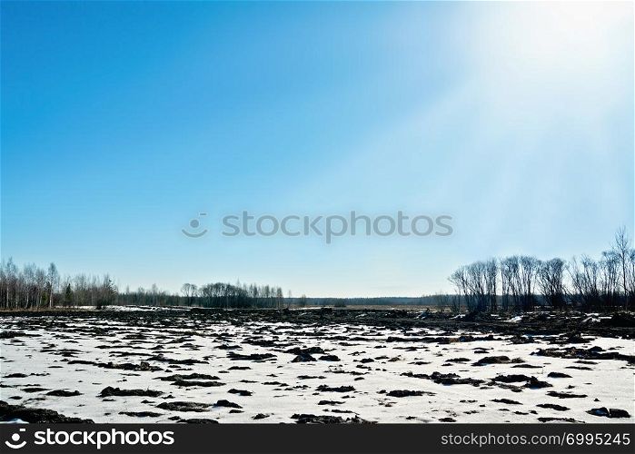 Landscape with melting snow on early spring land, blue sky, sun rays, trees and forest.