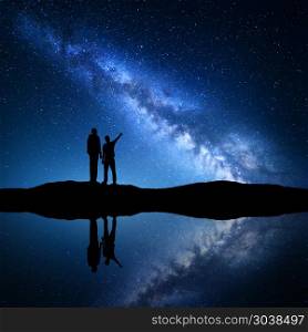 Landscape with Milky Way. Silhouette of a father and son. Milky Way with silhouette of a family. Father and a son who pointing finger in night starry sky on the mountain near the river with sky reflection in water. Night landscape. Space. Milky way with men