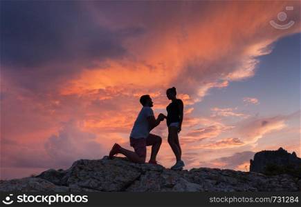 Landscape with silhouettes of a man making marriage proposal to his girlfriend on the mountain peak at colorful sunset. Silhouette of lovers. Couple. People, relationship