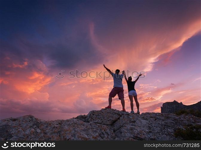 Landscape with silhouettes of a standing young man and woman on the mountain peak at colorful sunset. Man and girl on the background of beautiful cloudy sky. Silhouette of people. Couple. Travel