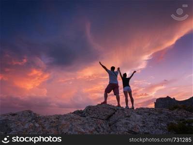 Landscape with silhouettes of a standing young man and woman on the mountain peak at colorful sunset. Man and girl on the background of beautiful cloudy sky. Silhouette of people. Couple. Travel